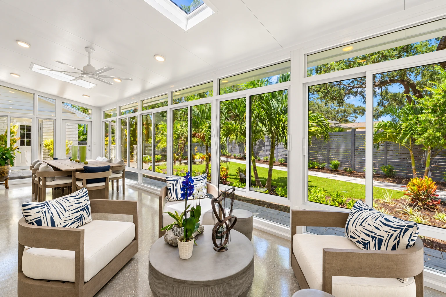 Bright sunroom with white-framed floor-to-ceiling windows, lounge chairs, a dining table, and a lush garden outside.