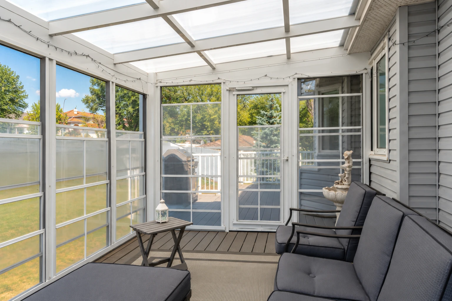 A bright enclosed patio has white-framed screen panels, a translucent roof, gray siding, and dark outdoor sofas.