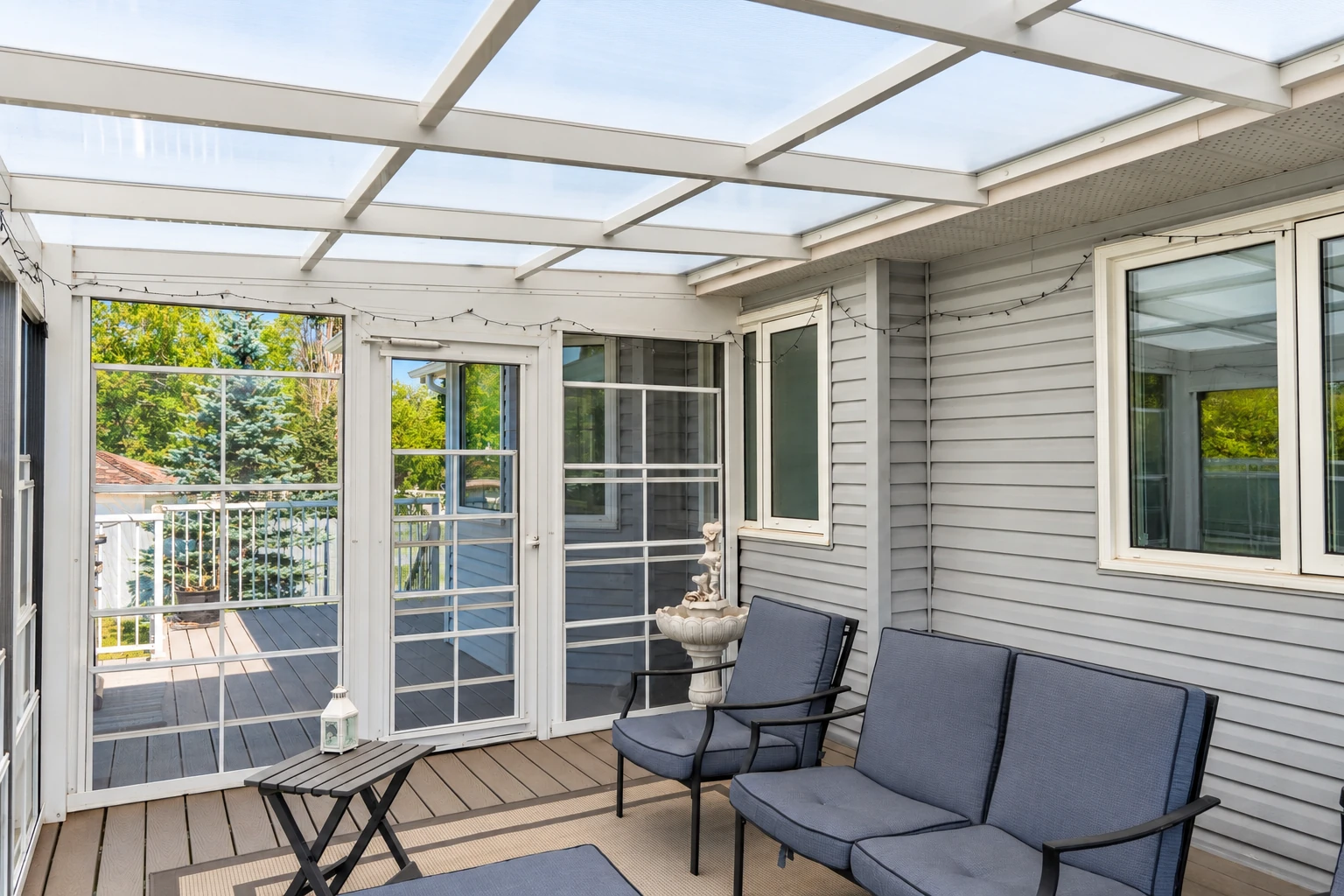 A screened porch with a translucent roof has gray siding, mesh panels, a door, and dark cushioned seating.