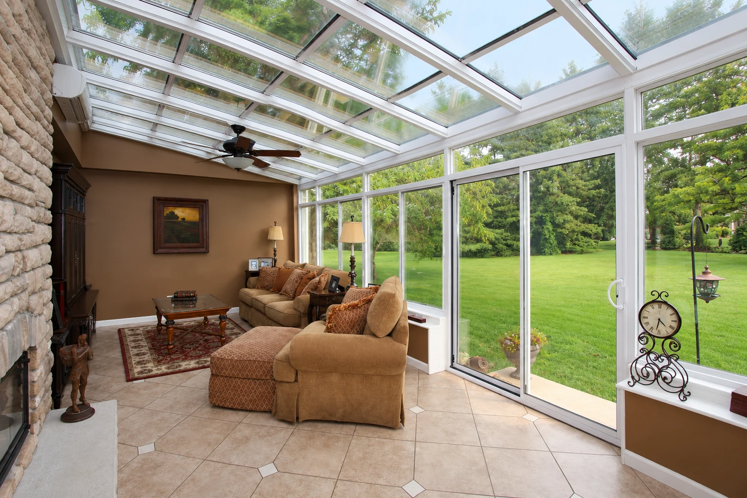 Solarium with a glass-paneled roof, white-framed windows, tiled floor, and tan seating overlooking a green lawn.