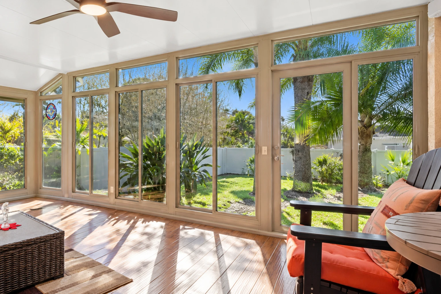 Bright sunroom with tall beige-framed windows, polished wood floors, and views of a palm-filled garden.