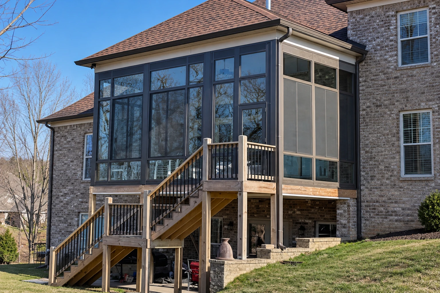 A dark-framed glass sunroom extends from a brick house, with a wooden deck and stairs beneath it on a clear day.