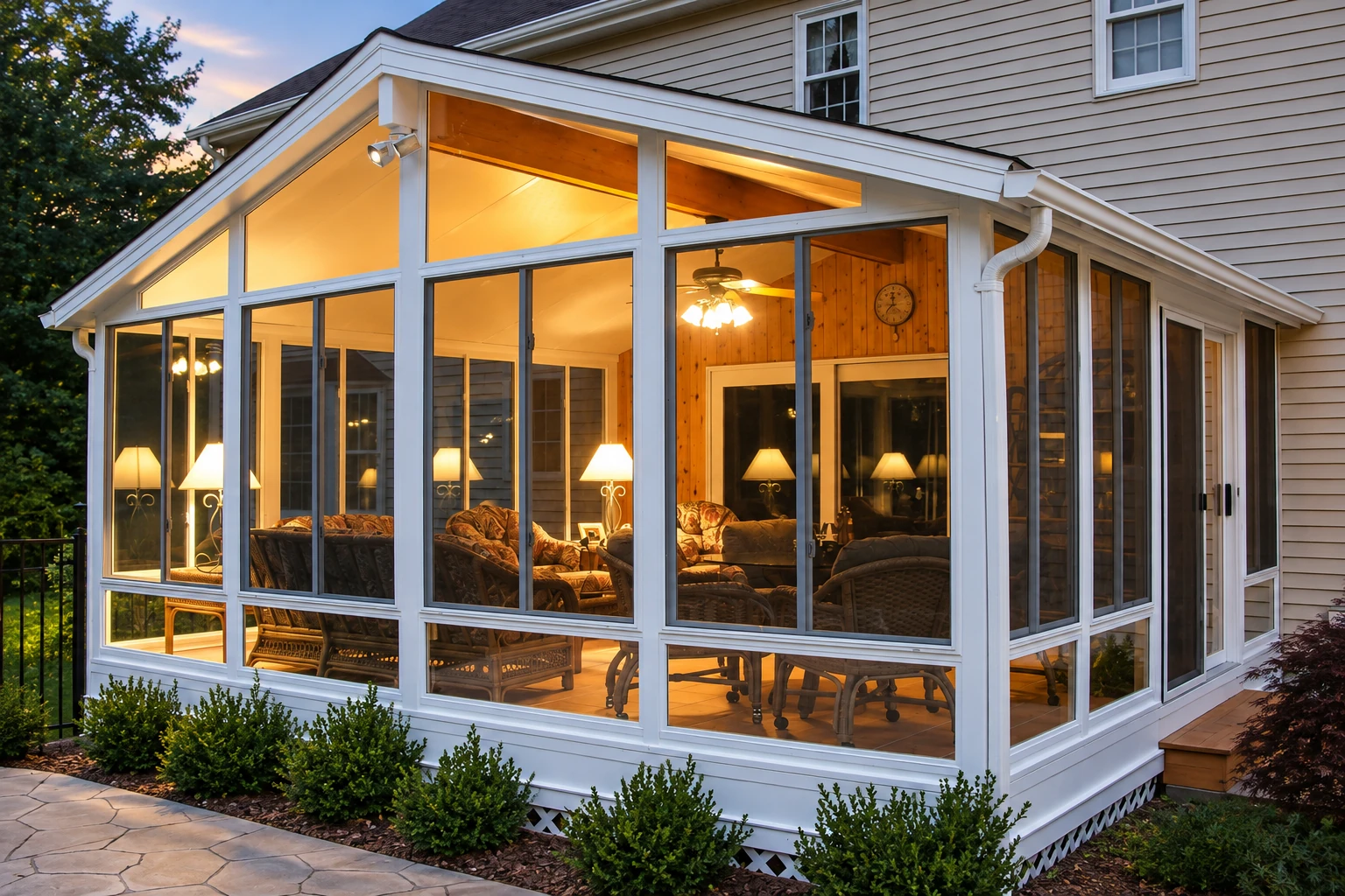 Three-season sunroom with white-framed glass walls, a sloped roof, and warmly lit wicker seating beside a house.