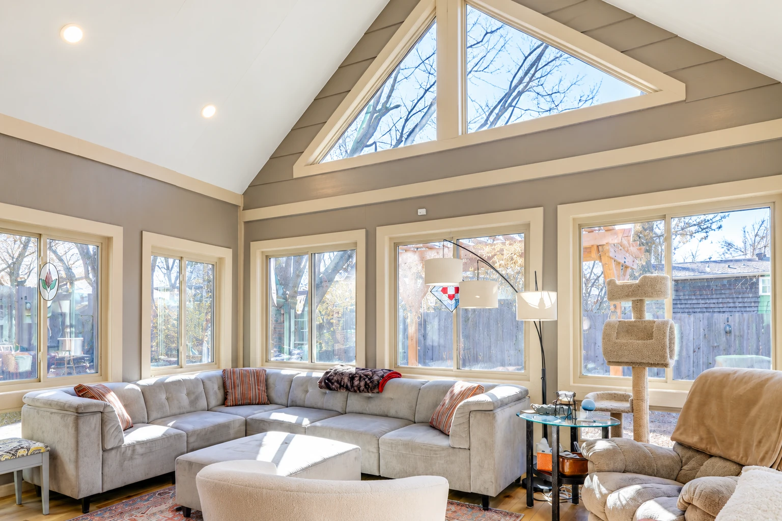 Bright sunroom with a high triangular window, wide windows, and a pale sectional sofa.